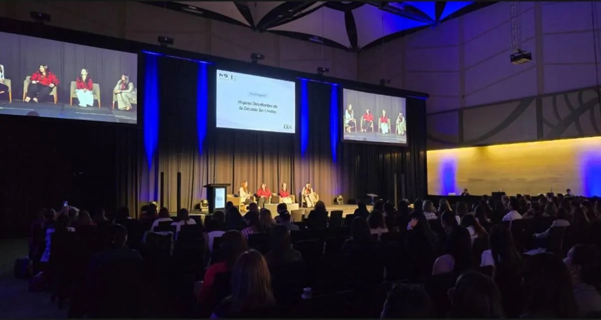 Tres mujeres sentadas en sillones en un escenario frente a una gran pantalla con texto de panel magistral