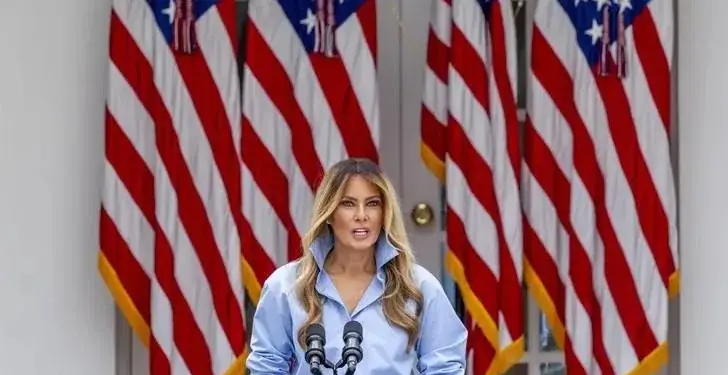 Una mujer con una camisa azul claro y un collar llamativo habla detrás de un atril con el sello presidencial de EE.UU., con varias banderas estadounidenses al fondo.