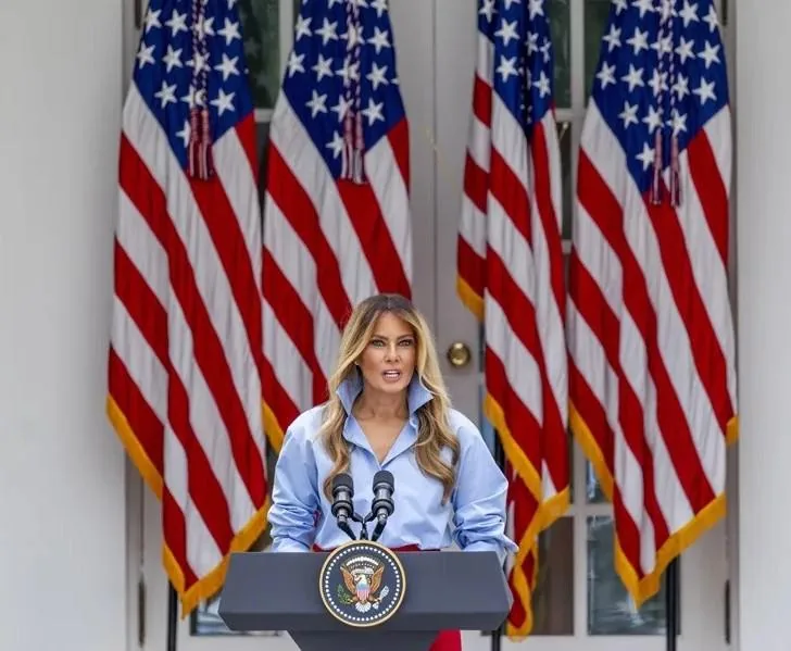 Una mujer con una camisa azul claro y un collar llamativo habla detrás de un atril con el sello presidencial de EE.UU., con varias banderas estadounidenses al fondo.