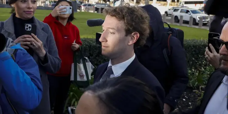 Mark Zuckerberg, vestido con traje y corbata, camina rodeado de personas, algunas sosteniendo teléfonos y cámaras, mientras es observado por un grupo de individuos, uno de ellos con gafas de sol.