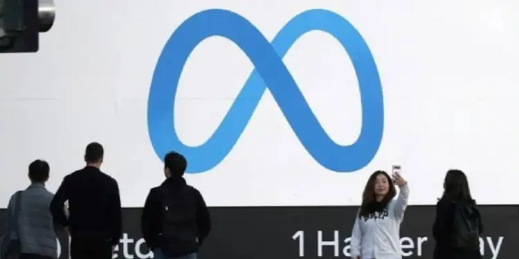 Personas de pie frente a una gran pantalla que muestra el logotipo azul en forma de infinito de Meta