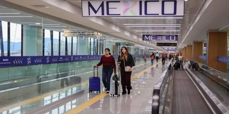 Dos mujeres caminan con maletas en un pasillo moderno de aeropuerto en México, con letreros de 'MEXICO' y vistas a una marina y montañas.