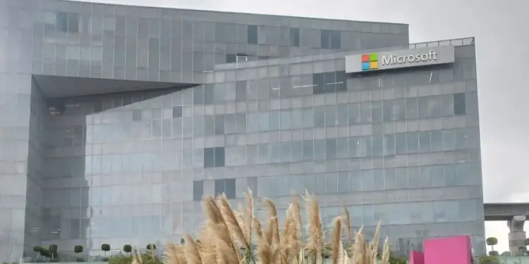 Vista exterior del moderno edificio de oficinas de Microsoft con fachada de cristal, rodeado de plantas altas y un cielo nublado.