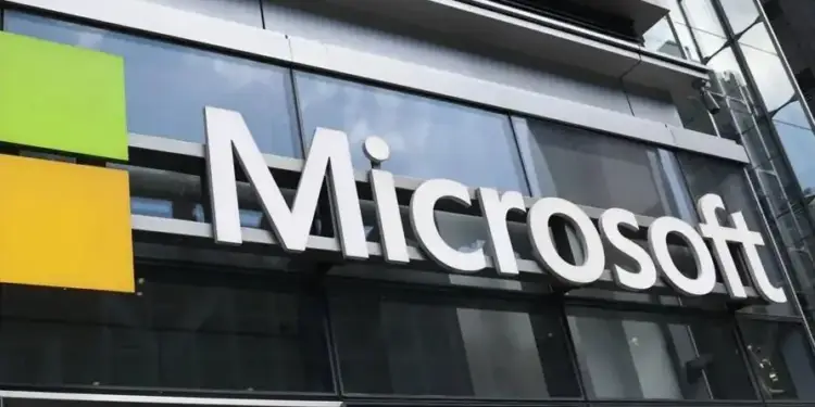 Primer plano del letrero de Microsoft en un edificio corporativo moderno con fachada de cristal. Se aprecian los cuadrados verde y amarillo del logo.