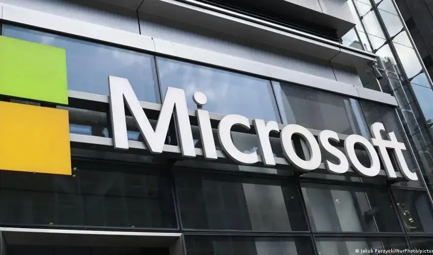 Primer plano del letrero de Microsoft en un edificio corporativo moderno con fachada de cristal. Se aprecian los cuadrados verde y amarillo del logo.