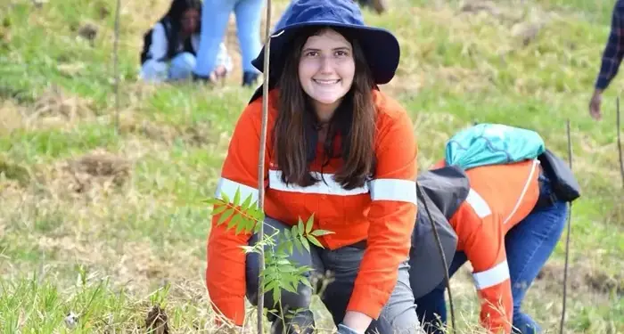 Mujer joven sonriente con sombrero y camisa naranja participando en una actividad de reforestación en un campo abierto.