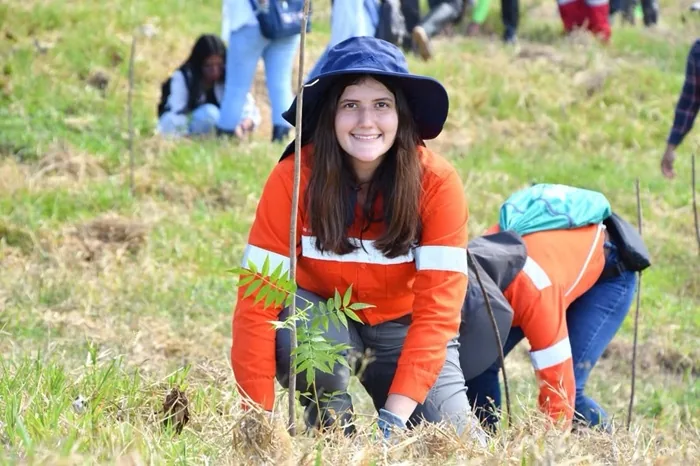 Mujer joven sonriente con sombrero y camisa naranja participando en una actividad de reforestación en un campo abierto.