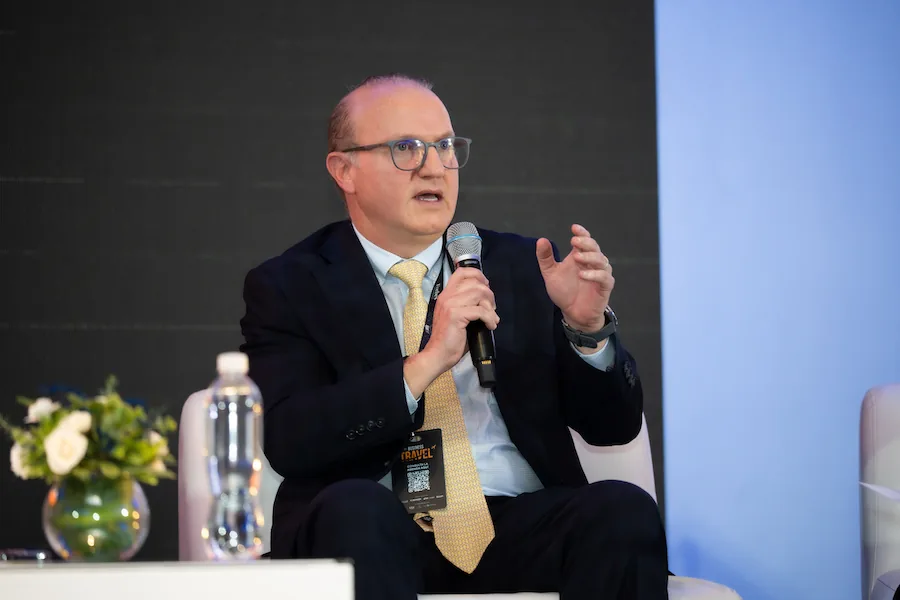 Hombre con traje, corbata y gafas hablando por micrófono en un evento de Business Travel.