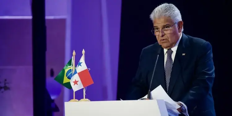 Hombre de negocios con traje y corbata hablando en un podio con banderas de Brasil y Panamá. Sostiene papeles y usa gafas, con fondo morado.