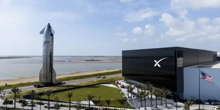 Cohete espacial metálico junto a un moderno edificio corporativo con logotipo y bandera de Estados Unidos al fondo.