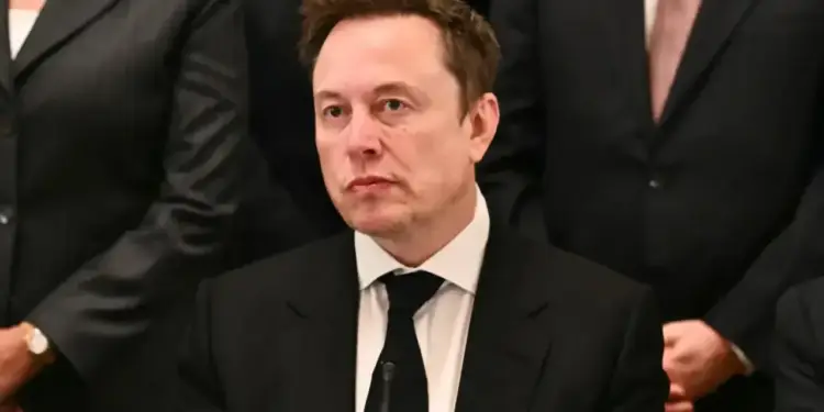 Elon Musk vistiendo traje formal y corbata negra en un entorno de negocios con otras personas de fondo.
