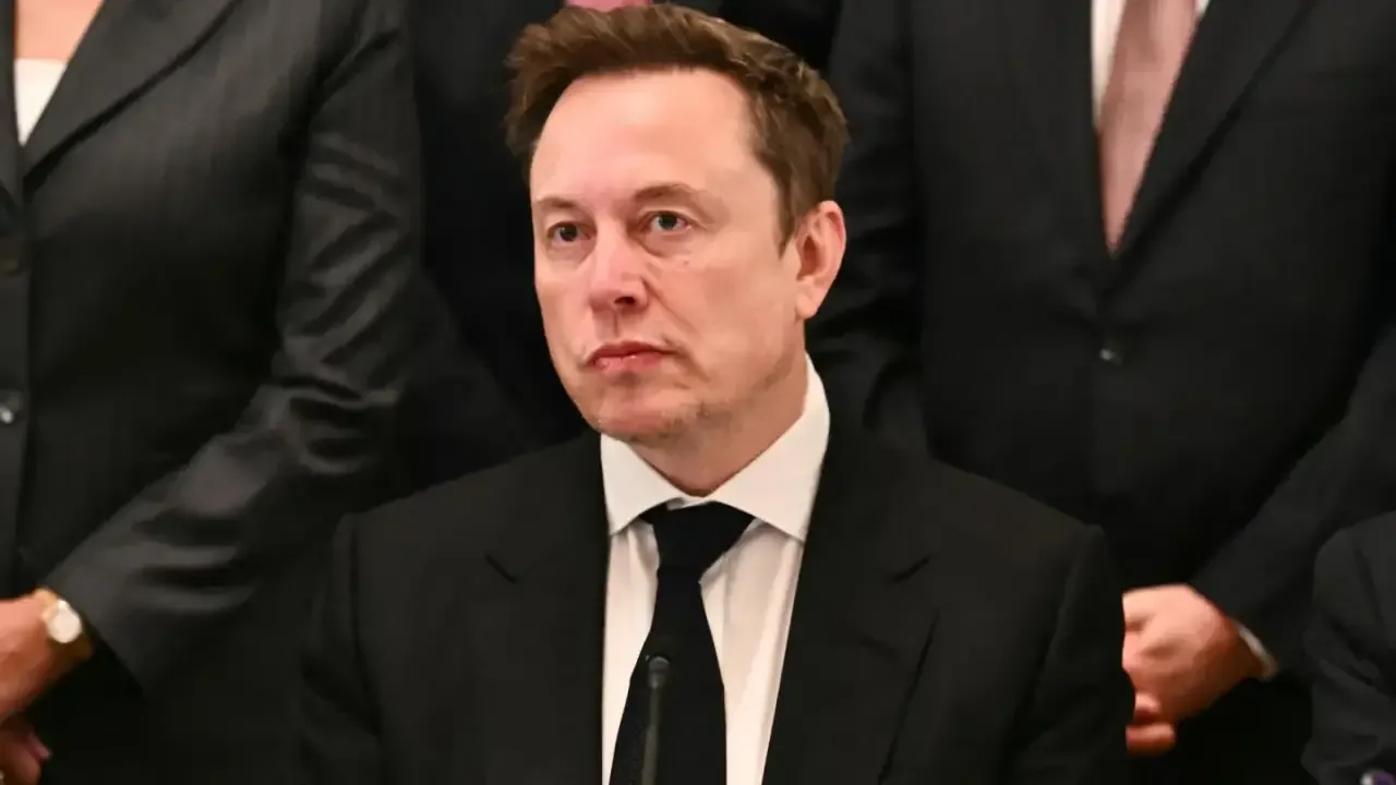 Elon Musk vistiendo traje formal y corbata negra en un entorno de negocios con otras personas de fondo.