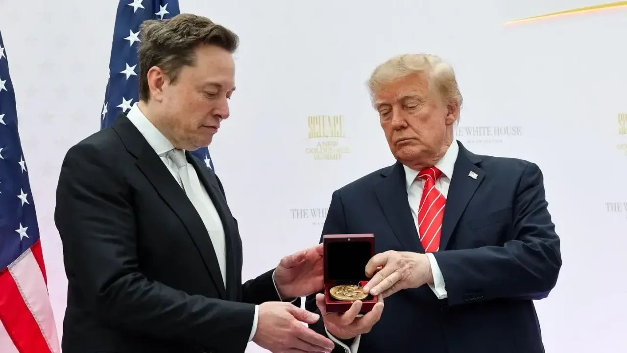 Dos hombres con traje formal sostienen una pequeña caja roja con una medalla dorada frente a banderas de Estados Unidos.
