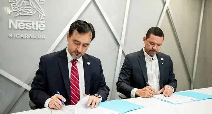 Dos hombres con traje firmando documentos en una mesa frente a un logotipo corporativo en la pared.