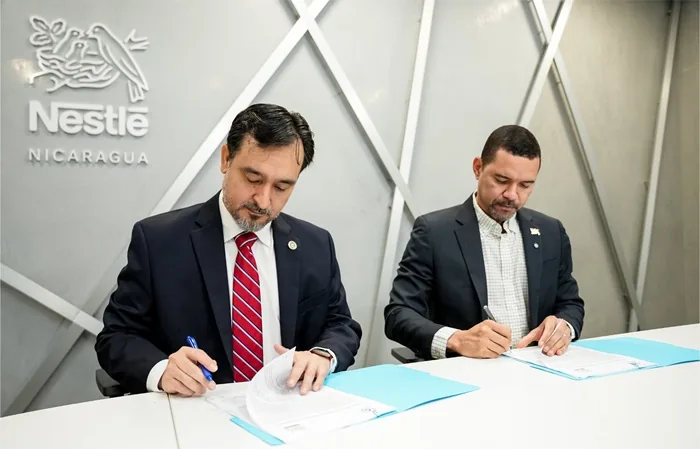 Dos hombres con traje firmando documentos en una mesa frente a un logotipo corporativo en la pared.