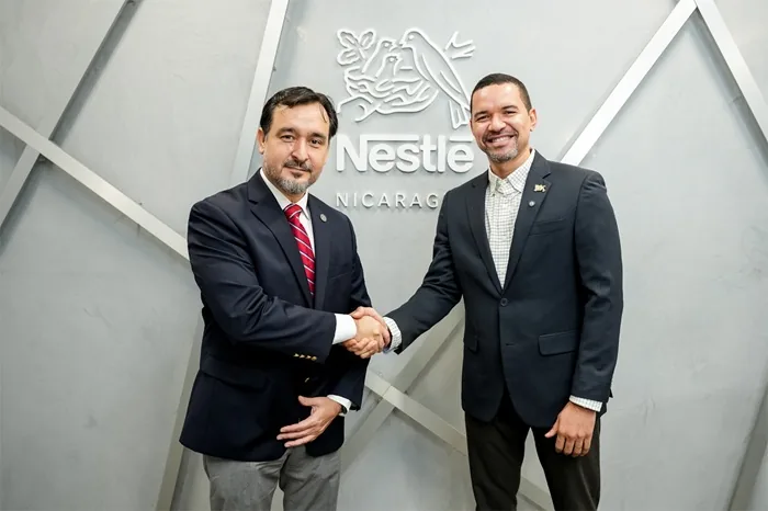 Dos hombres de negocios con traje formal estrechando las manos frente al logo de Nestle Nicaragua en una oficina moderna.