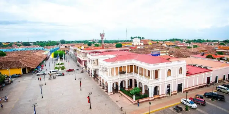 Vista aérea de una plaza central con edificios coloniales de techos rojos y blancos, personas caminando, vehículos estacionados y vegetación. El cielo está nublado.