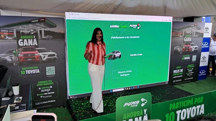 Mujer presentando un sorteo empresarial frente a una gran pantalla digital verde con texto promocional y vehículos.