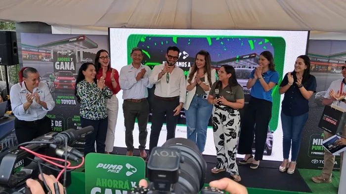 Grupo de personas aplaudiendo en un evento corporativo frente a una pantalla con un expositor con micrófono.
