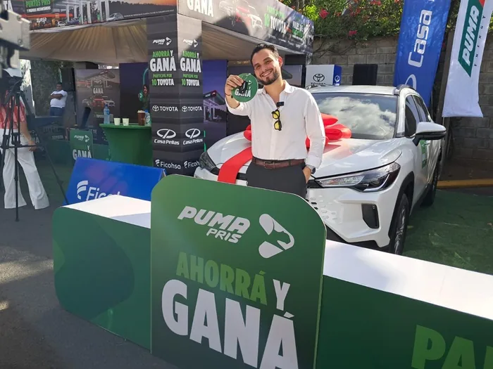Hombre sonriente sosteniendo la llave de un auto blanco nuevo junto a un stand promocional con elementos corporativos.