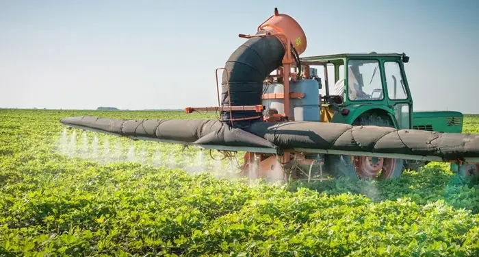 Tractor verde equipado con sistema de fumigación trabajando en un campo de cultivo verde bajo el sol.