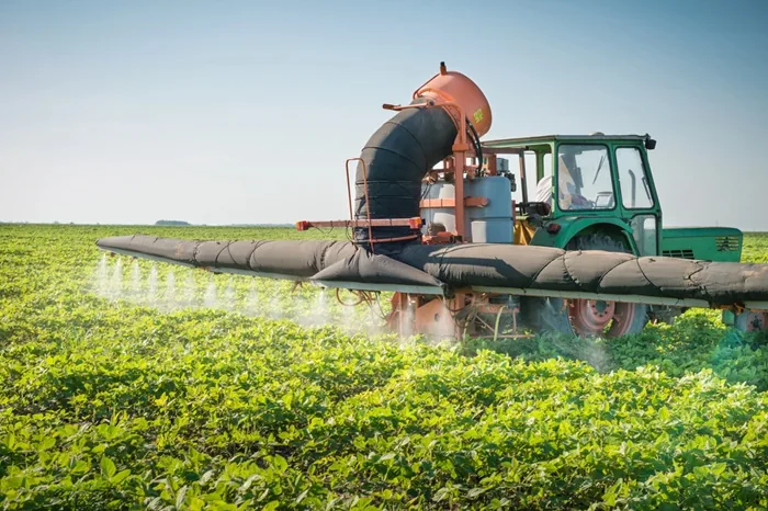 Tractor verde equipado con sistema de fumigación trabajando en un campo de cultivo verde bajo el sol.