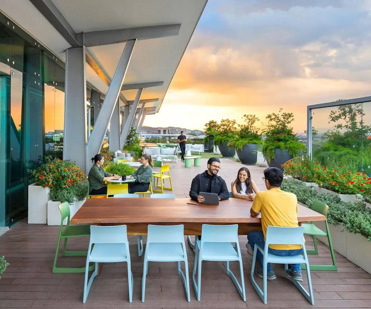 Un grupo de jóvenes profesionales interactuando en una terraza moderna con mobiliario de diseño, plantas y vistas al atardecer, sugiriendo un ambiente de colaboración y bienestar.