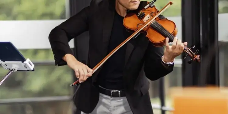 Hombre joven con traje negro y pantalón gris tocando el violín con un micrófono, frente a una tableta en un soporte.