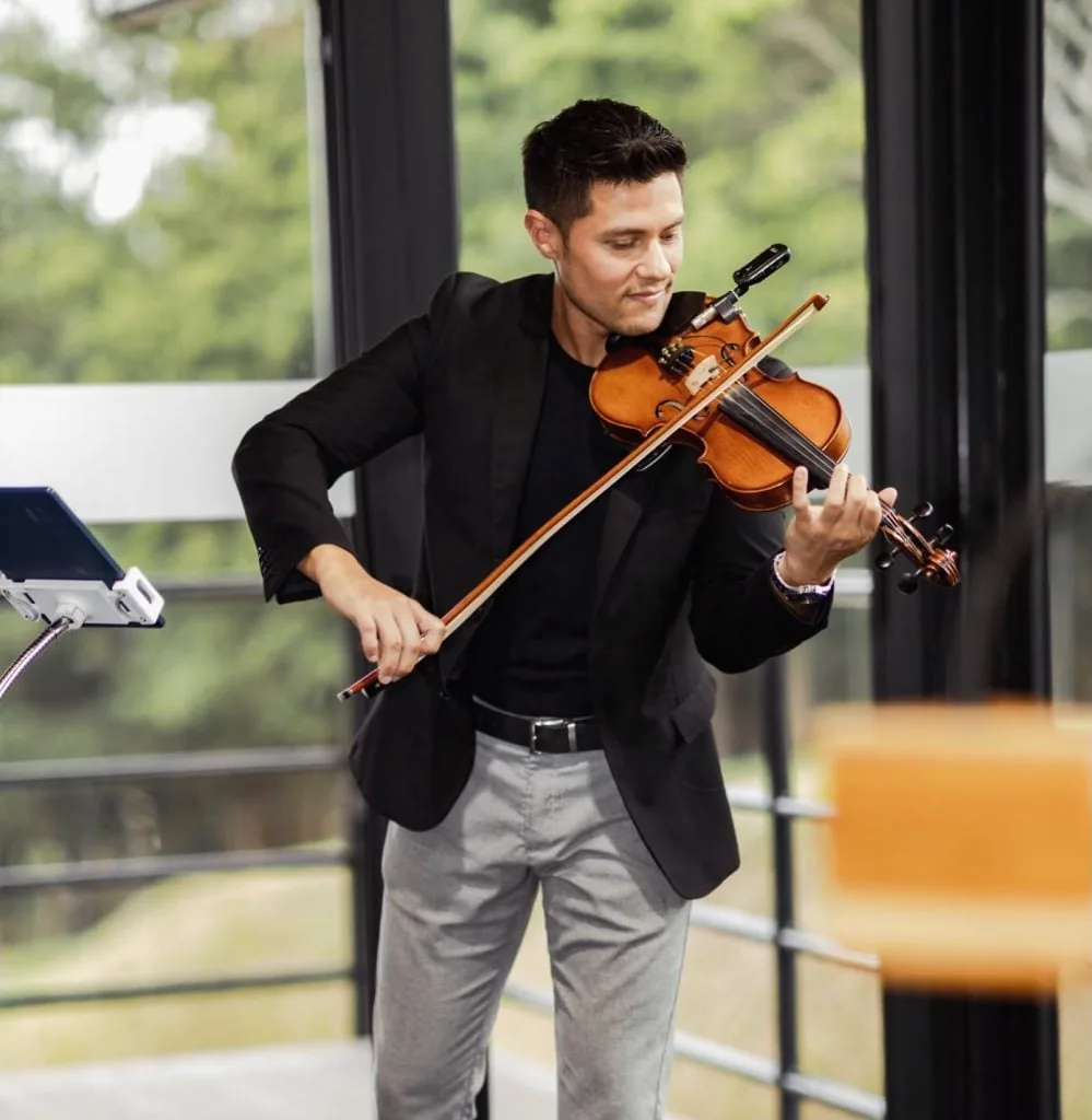 Hombre joven con traje negro y pantalón gris tocando el violín con un micrófono, frente a una tableta en un soporte.
