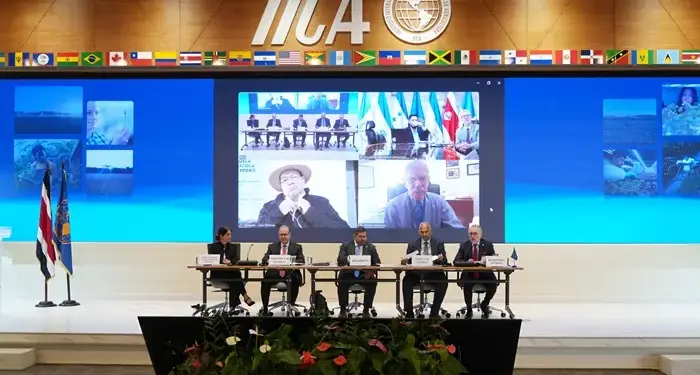 Cinco personas sentadas en una mesa directiva frente a una gran pantalla con videoconferencia y el logotipo de IICA.
