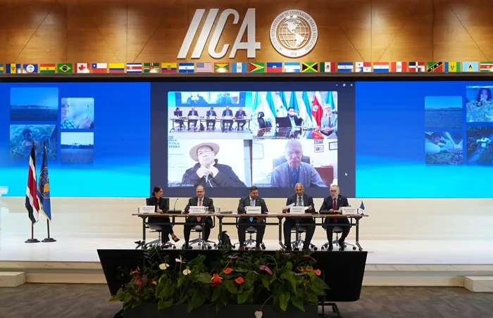 Cinco personas sentadas en una mesa directiva frente a una gran pantalla con videoconferencia y el logotipo de IICA.