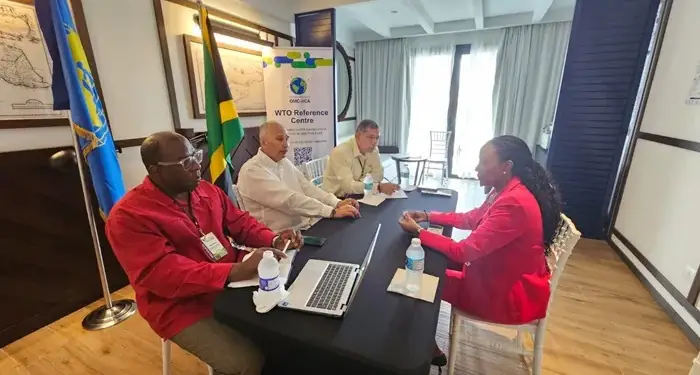 Cuatro profesionales sentados alrededor de una mesa negra en una reunión de negocios junto a banderas y un banner de la OMC.