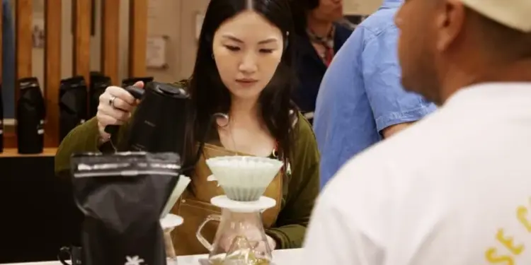 Una mujer joven barista preparando café filtrado con una tetera negra y un cono goteador en un mostrador.
