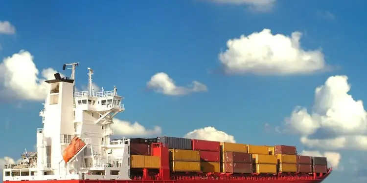 Gran barco portacontenedores rojo y blanco navegando en el mar bajo un cielo azul con nubes blancas.