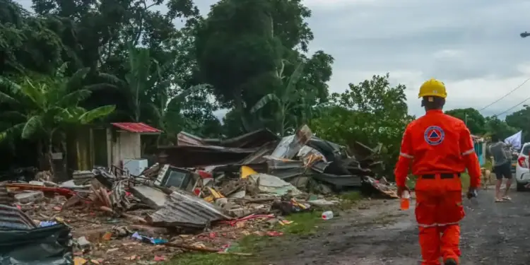 Trabajador de rescate camina entre escombros de edificios destruidos tras un desastre, con vegetación tropical de fondo.