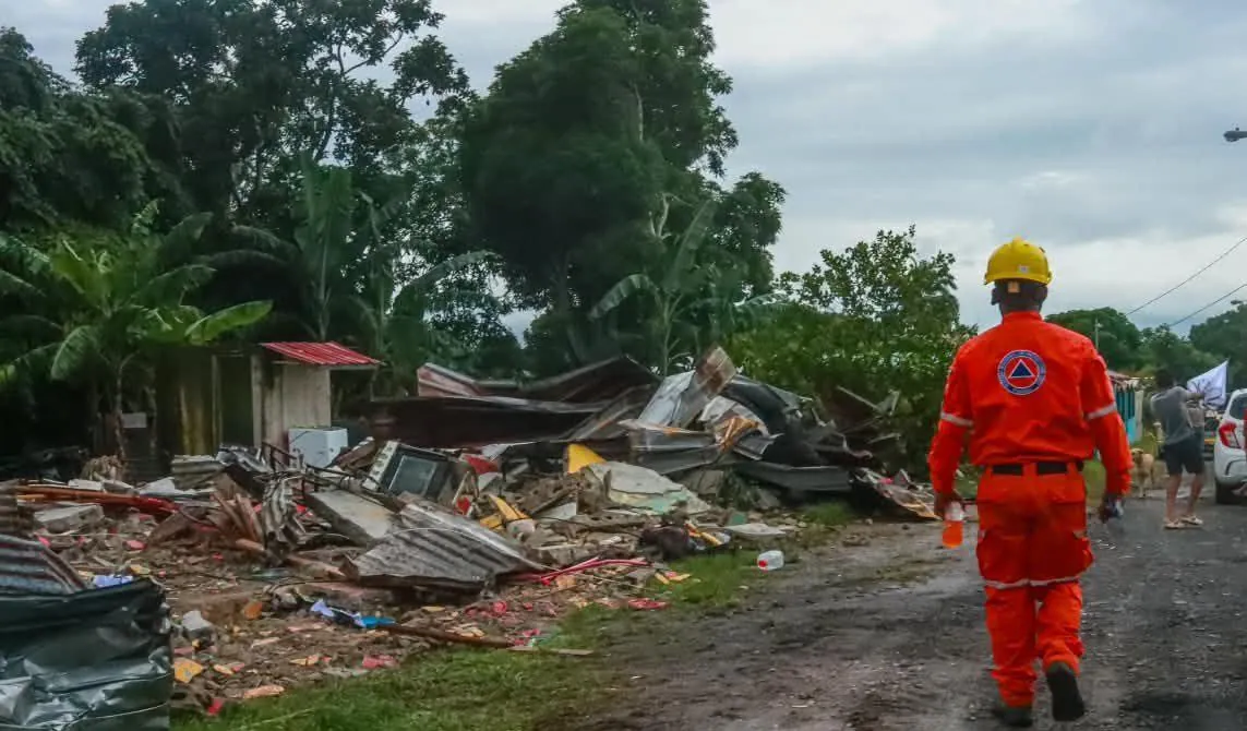 Trabajador de rescate camina entre escombros de edificios destruidos tras un desastre, con vegetación tropical de fondo.