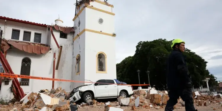 Edificio blanco con torre dañada y escombros en la calle, junto a una camioneta blanca y un rescatista con casco amarillo.
