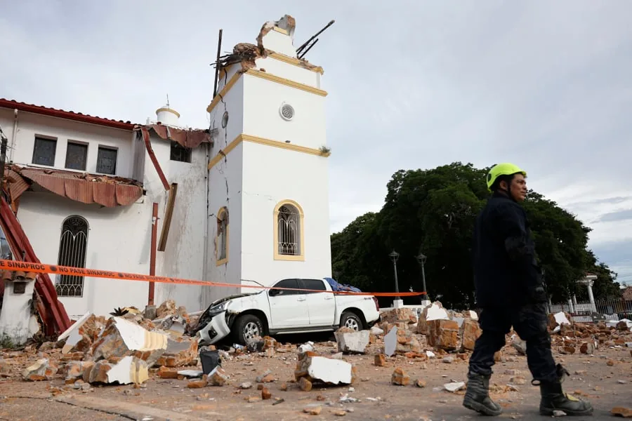 Edificio blanco con torre dañada y escombros en la calle, junto a una camioneta blanca y un rescatista con casco amarillo.