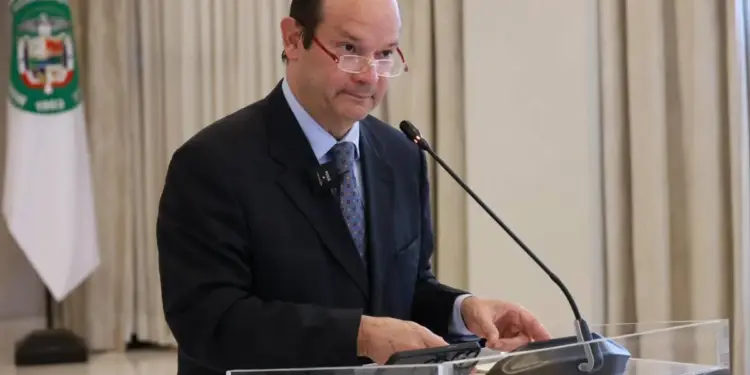 Orador masculino con traje oscuro, corbata y gafas, presentando en un podio de acrílico con micrófono. Sostiene un dispositivo móvil, con una bandera institucional al fondo.