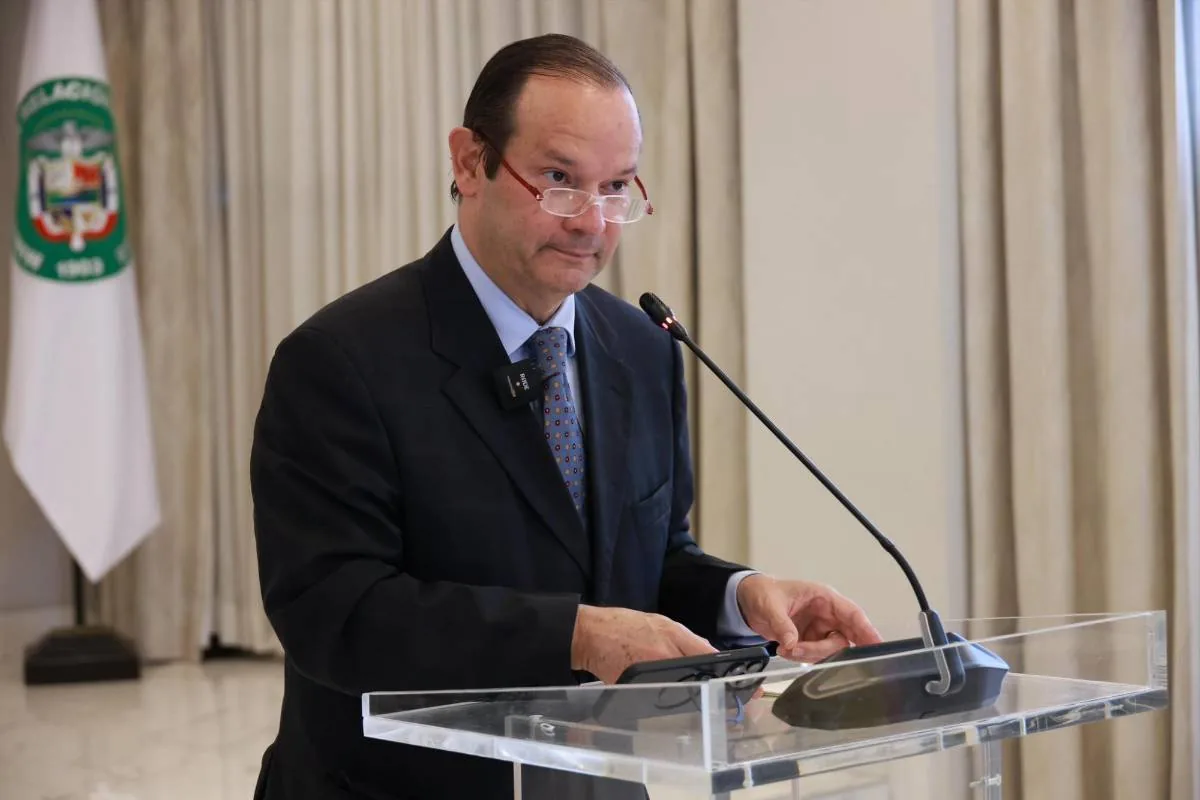 Orador masculino con traje oscuro, corbata y gafas, presentando en un podio de acrílico con micrófono. Sostiene un dispositivo móvil, con una bandera institucional al fondo.