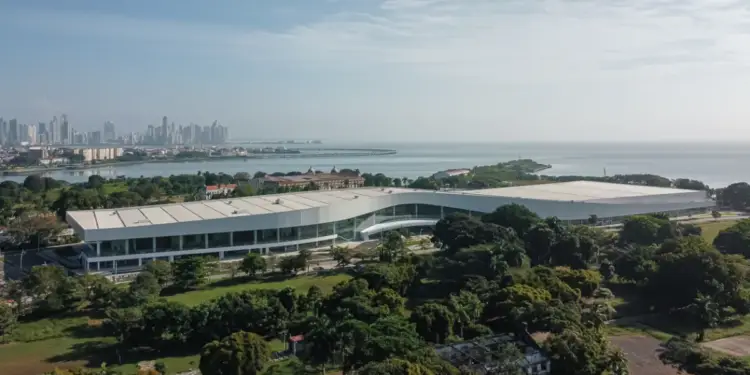 Vista aérea de un moderno centro de convenciones blanco rodeado de vegetación frente al mar y la ciudad.