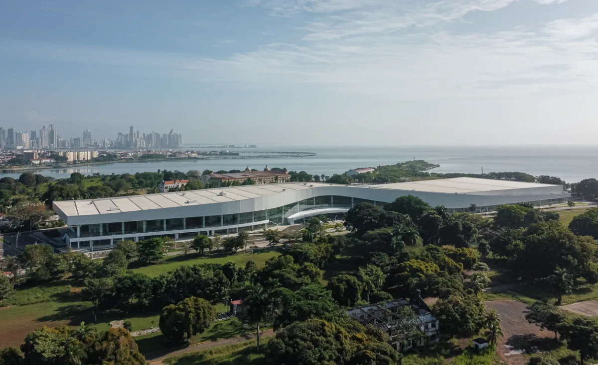 Vista aérea de un moderno centro de convenciones blanco rodeado de vegetación frente al mar y la ciudad.