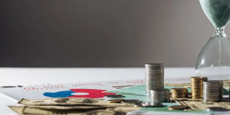 Reloj de arena junto a pilas de monedas y billetes sobre documentos financieros y gráficos.