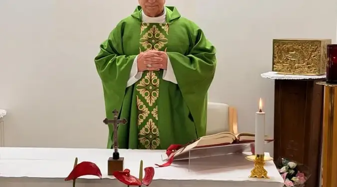 Un sacerdote con vestimenta litúrgica verde y blanca, con las manos juntas, frente a un altar con un crucifijo, un libro abierto y flores rojas y blancas.