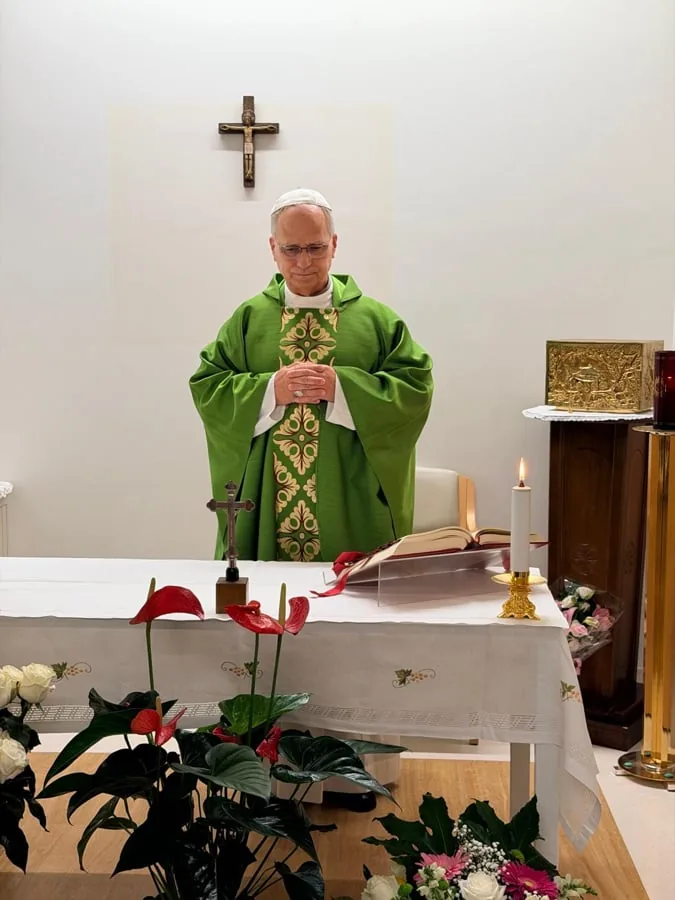 Un sacerdote con vestimenta litúrgica verde y blanca, con las manos juntas, frente a un altar con un crucifijo, un libro abierto y flores rojas y blancas.