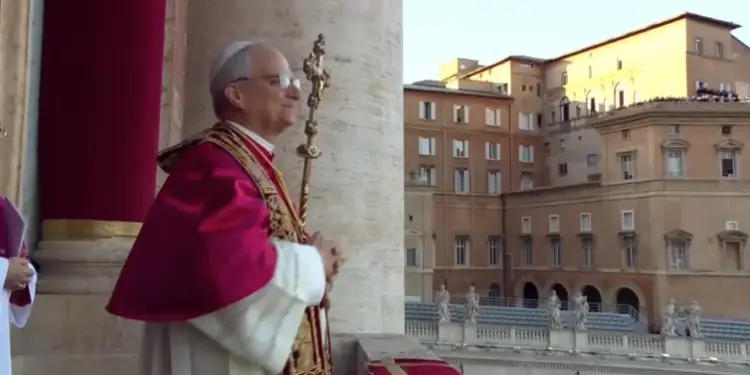 Sacerdote con vestimenta formal y báculo en un balcón junto a grandes columnas y edificios históricos al fondo.