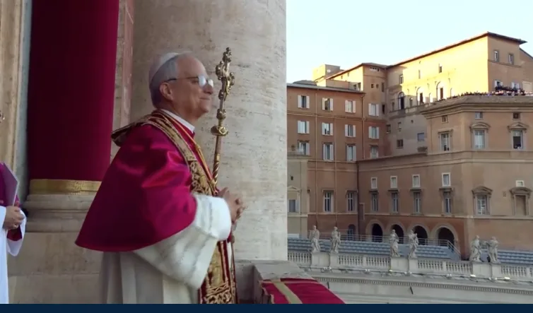 Sacerdote con vestimenta formal y báculo en un balcón junto a grandes columnas y edificios históricos al fondo.