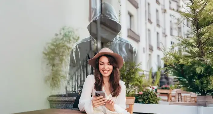 Mujer sonriente usando su móvil en una terraza, con una figura encapuchada translúcida detrás, simbolizando riesgos de ciberseguridad.