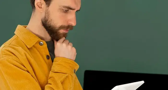 Hombre con barba y camisa amarilla mirando pensativo una tablet digital en su mano