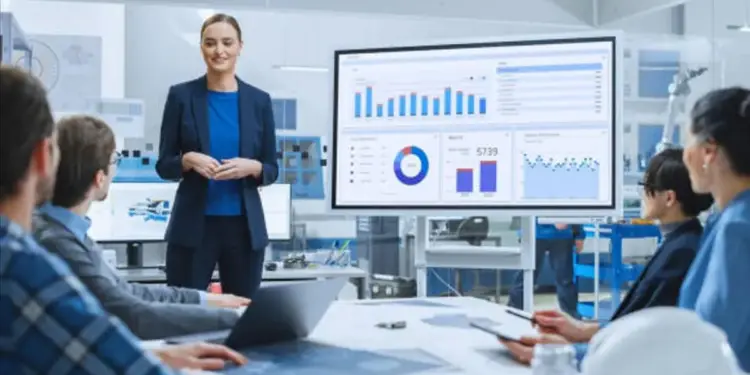 Mujer profesional presenta análisis de datos en pantalla grande a un equipo en reunión. Gráficos y cuadros de mando visibles. Entorno de oficina.
